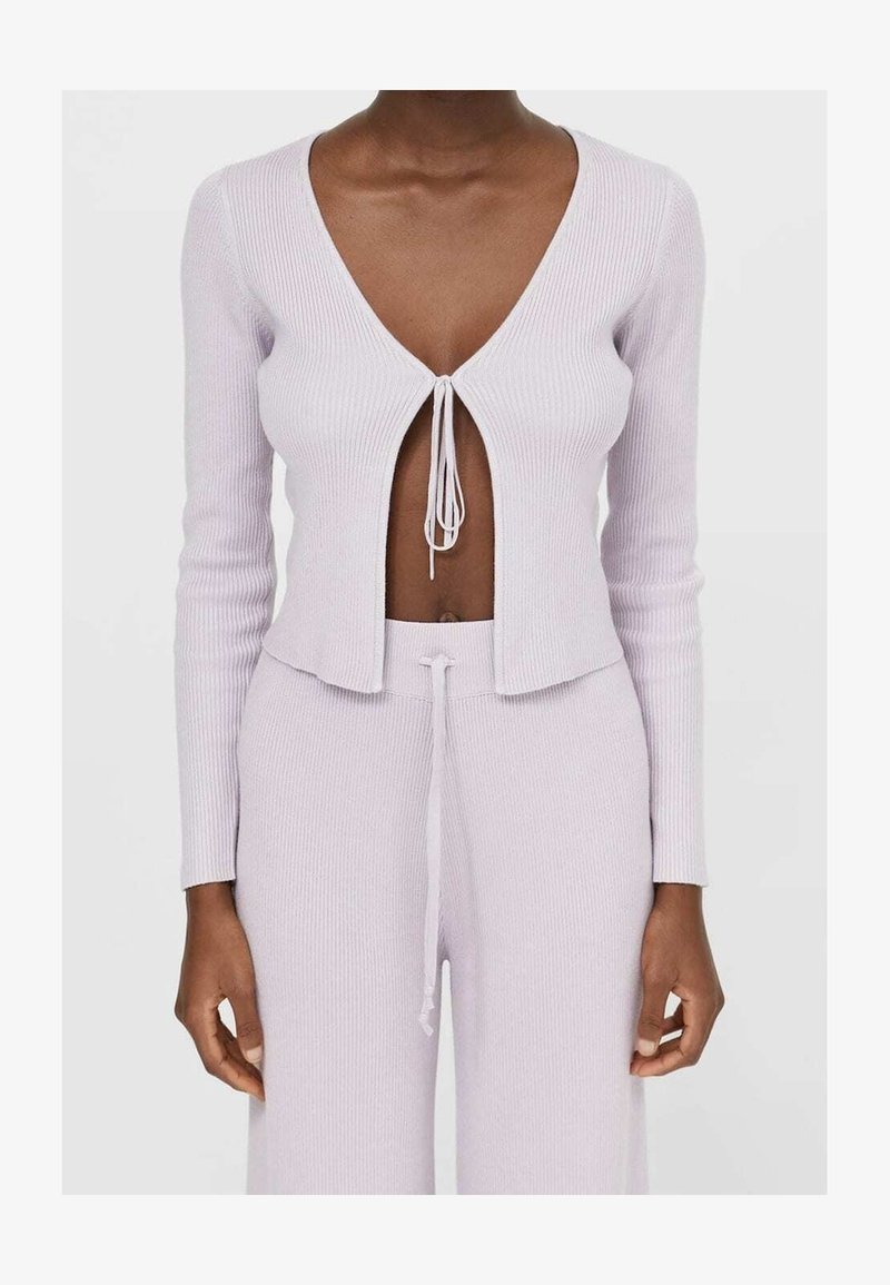 Cardigan court en maille côtelée lilas avec manches longues, encolure en V profonde et fermeture à nouer à l'avant. Associé à un pantalon assorti.