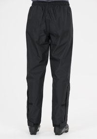 Person iført sorte vandtætte bukser med elastisk talje og justerbare ankelstropper, stående mod en hvid baggrund.