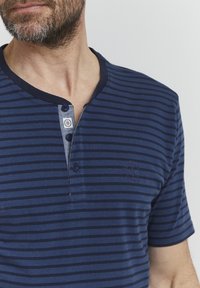 Kleita ar īsām piedurknēm, tumši zila ar svītrām, ar trīs pogu henley kaklu un nelielu logo detaļu kreisajā krūtī. Mīksta kokvilnas audums.