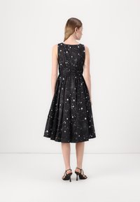 Black dress with silver star pattern, sleeveless design, flared skirt, back waistband detail, with model wearing black heels.