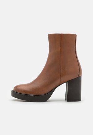 Stivaletto alla caviglia in pelle marrone con punta arrotondata, tacco alto a blocco e texture liscia. Presenta cuciture minimali e una silhouette elegante.