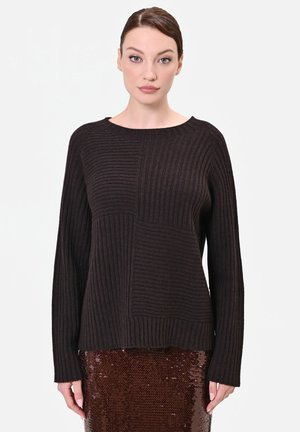 Donna con maglione a coste marrone scuro e gonna marrone con paillettes, in piedi davanti a uno sfondo bianco semplice.