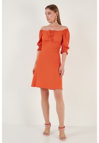 Orange off-shoulder dress with puff sleeves, ruched bodice, and a flared hemline. Worn with multicolored high-heeled sandals and statement earrings.