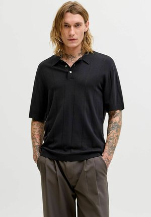 Ung mann med skulderlangt blondt hår, tatoverte armer og nakke, iført svart kortermet poloshirt og grå plisserte bukser, hendene i lommene.