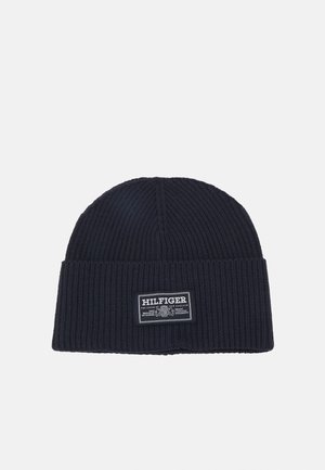 Marineblauwe gebreide beanie met geribbeld textuur en een opgerolde manche. Voorzien van een wit etiket met de merknaam en het logo aan de voorkant.