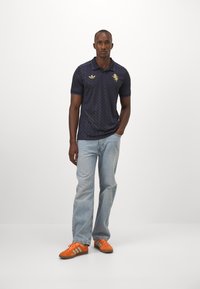 Marineblaues Poloshirt mit strukturiertem Muster, goldenem Löwenlogo, kombiniert mit hellblauen Jeans und orangefarbenen Wildledersneakern mit türkisfarbenen Streifen.