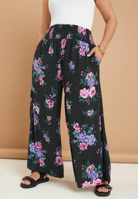 Svarta blommiga byxor med vida ben och smockad midja, med rosa, lila och gröna blommor. Fickor ingår, stiliserade med platta svarta sandaler.