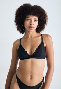 Musta pitsinen bralette, jossa on ohuet säädettävät olkaimet, aaltoilevat reunat ja pehmeä kangas; yhdistettynä vastaaviin mustiin pitsisiin alushousuihin.