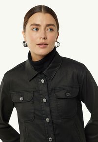 Czarna dżinsowa kurtka z dwiema kieszeniami na piersi, srebrnymi guzikami i kołnierzem. Noszona na czarnym golfie. Gładka faktura.