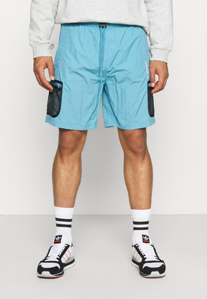 Man wearing light blue shorts with black mesh pockets, white Adidas socks with black stripes, and black-and-white Adidas sneakers.