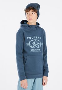 Felpa blu navy con tasca a marsupio, caratterizzata da una stampa grafica con montagne e occhiali da sci. Indossata con un berretto a coste abbinato.