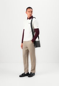 Chemise blanche à manches courtes avec logo, chemise bordeaux à manches longues en dessous, pantalon beige, chaussures en cuir noir et un sac messager noir.