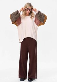 Oversized kremowa bluzka z wzorzystymi brązowymi i rdzawymi bufkami, w zestawie z ciemnobrązowymi spodniami o szerokich nogawkach z białymi paskami po bokach.