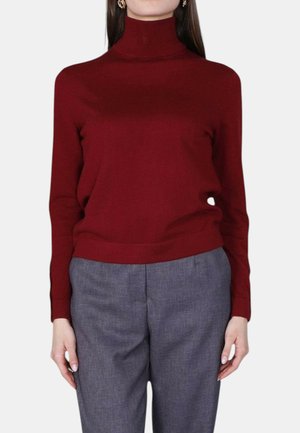 Donna che indossa un dolcevita aderente a maniche lunghe color borgogna e pantaloni grigi su misura, su uno sfondo chiaro e uniforme.