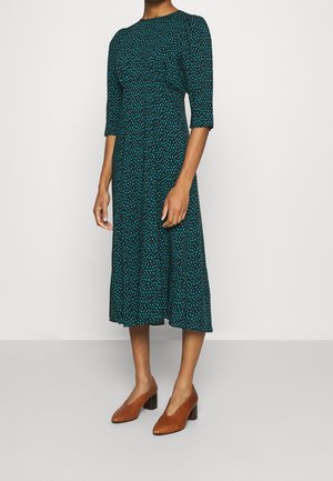 Vrouw die een donkergroene midi-jurk met blauwe vlekken en driekwart mouwen draagt, gecombineerd met bruine schoenen met blokhak.