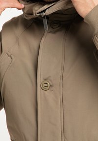 Giacca isolante marrone con colletto alto, chiusura con bottoni e zip, e una trama di tessuto che mostra una leggera lucentezza. Presenta tasche laterali.