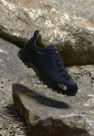 Donkerblauwe en zwarte Gore-Tex wandelschoen met Vibram-zool van The North Face die boven rotsachtig terrein zweeft.