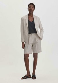 Lichtbeige blazer over een donkergrijze V-hals top, gecombineerd met bijpassende lichtbeige shorts. Het model draagt zwarte sandalen met een geknoopte band.