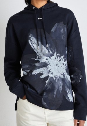Personne portant un sweat à capuche foncé avec un grand motif fleur gris et blanc et le mot BOSS près de l'encolure, associé à un pantalon clair.