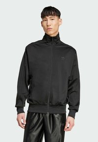Zwarte zip-up sportjack gemaakt van een gladde stof, met een rechte kraag, drie strepen langs de mouwen en een klein logo op de borst.