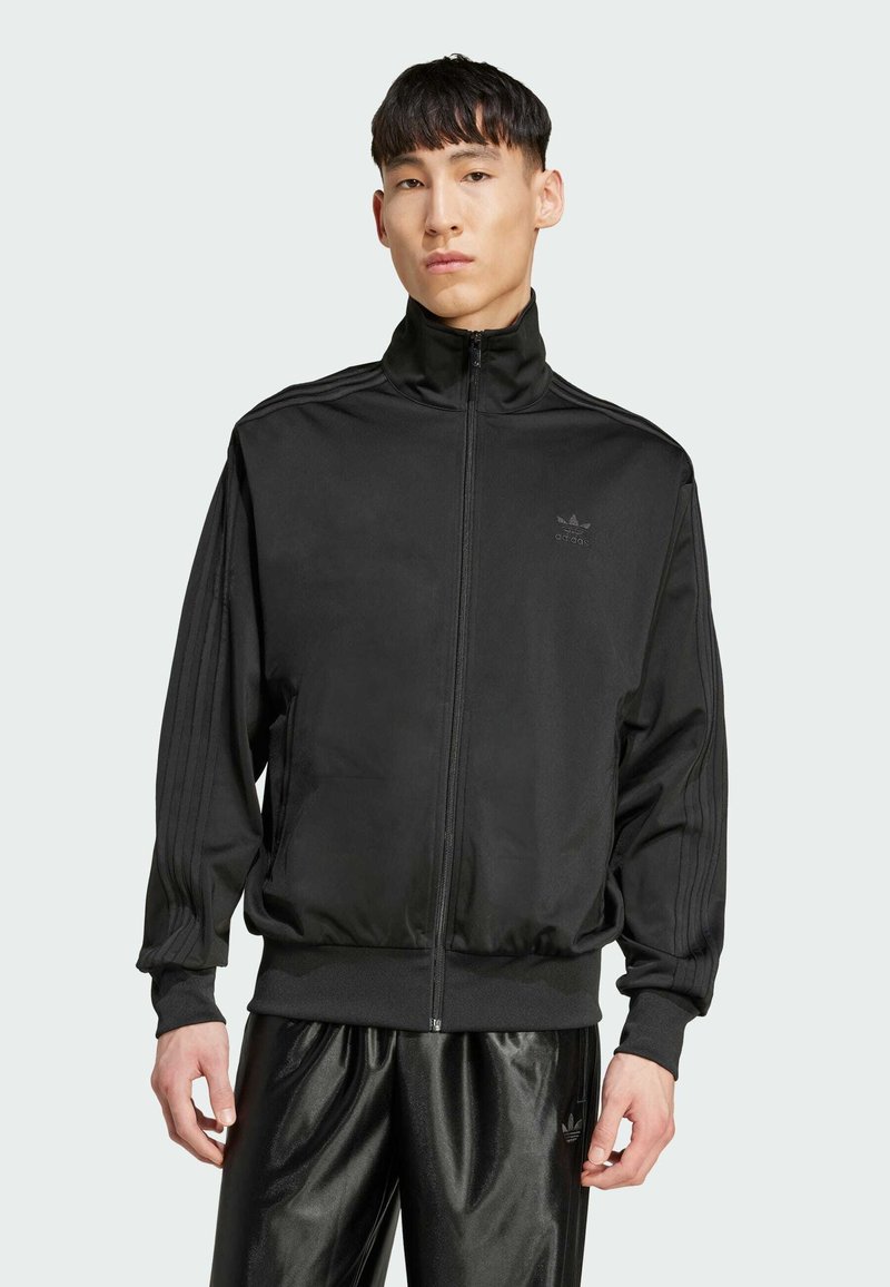 Zwarte zip-up sportjack gemaakt van een gladde stof, met een rechte kraag, drie strepen langs de mouwen en een klein logo op de borst.