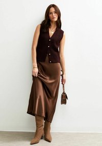 Woman wearing sleeveless dark brown knit vest, silky brown skirt, tan pointed boots, holding a small brown snakeskin handbag against white wall.