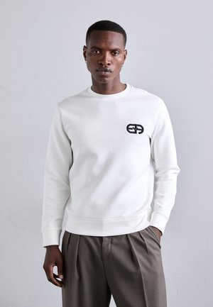 Witte sweatshirt van zacht materiaal met een zwart logo op de borst, ronde hals en ribgebreide manchetten. In combinatie met grijze broeken.