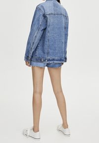 Henkilö, joka pitää yllään sinistä farkkutakkia ja siihen sopivia farkkuhousuja, seisoo selin kameraan, jalassaan valkoiset lenkkarit yksinkertaisella taustalla.