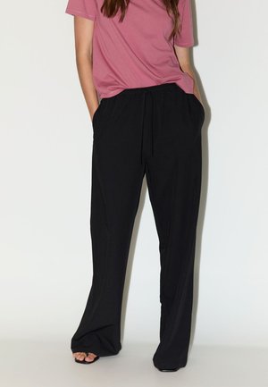 Personne portant une chemise rose à manches courtes et un pantalon noir ample, les mains dans les poches, debout devant un fond blanc uni.