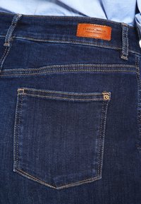 Jeans de mezclilla azul oscuro con un bolsillo trasero, costuras en contraste y un parche de cuero marrón con el logo. Textura suave, diseño ajustado y estilo de talle medio.
