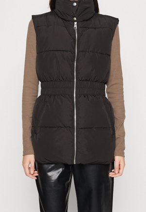 Schwarze gesteppte ärmellose Weste mit Frontreißverschluss und hohem Stehkragen mit Druckknöpfen, getragen über einem braunen Langarmshirt und schwarzen Lederhosen.
