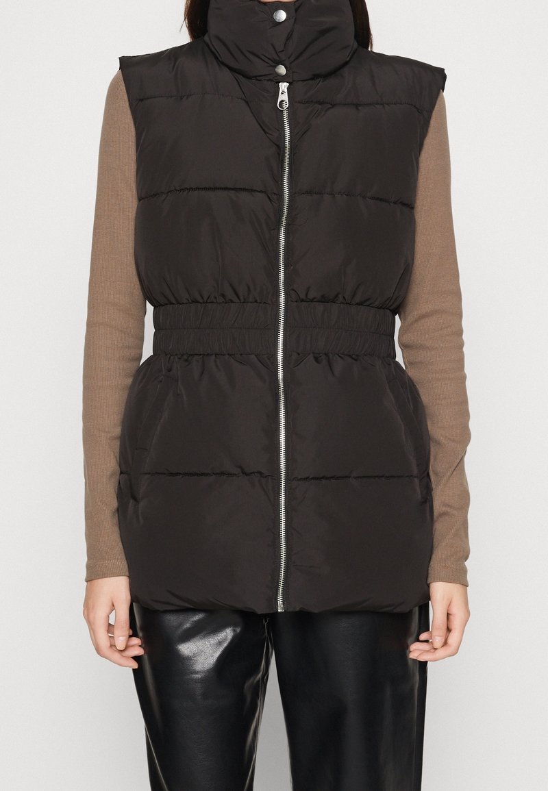 Veste noire matelassée sans manches avec fermeture éclair devant et col montant à boutons-pression, portée sur un top marron à manches longues et un pantalon en cuir noir.