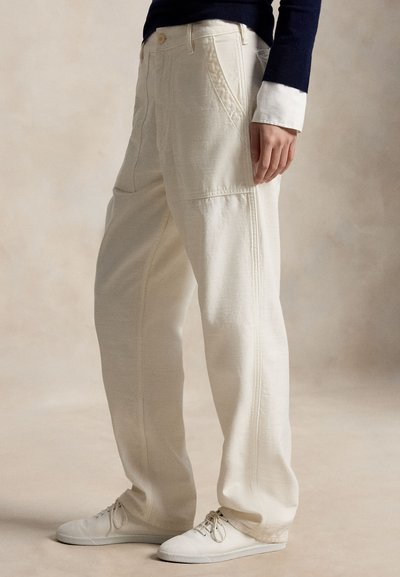 Persoană purtând pantaloni albi largi cu buzunare laterale și adidași albi, stând în fața unui fundal neutru.