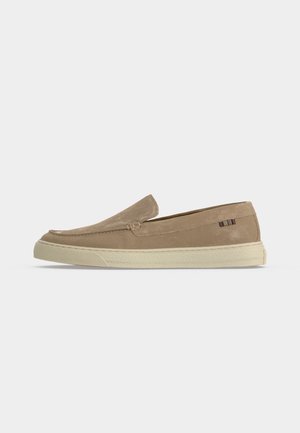 Beigen värinen mokkanahkainen slip-on-mokkasiini, jossa valkoinen kumipohja ja pieni monivärinen kangasmerkki kantapään sivulla.