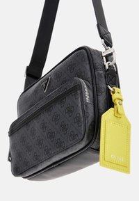 Borsa a tracolla nera in materiale sintetico con finitura testurizzata, dotata di una tasca con zip, accenti del logo e un'etichetta gialla. Include una tracolla regolabile.