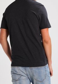 Persona che indossa una maglietta a maniche corte grigio scuro e jeans di denim blu chiaro, voltata di spalle su uno sfondo neutro.