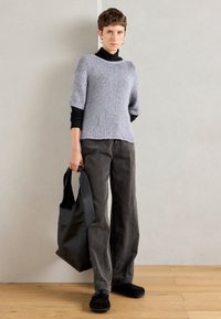 Person wearing a grey short-sleeve sweater over black long sleeves, dark wide-leg jeans, black shoes, and holding a large black tote bag.