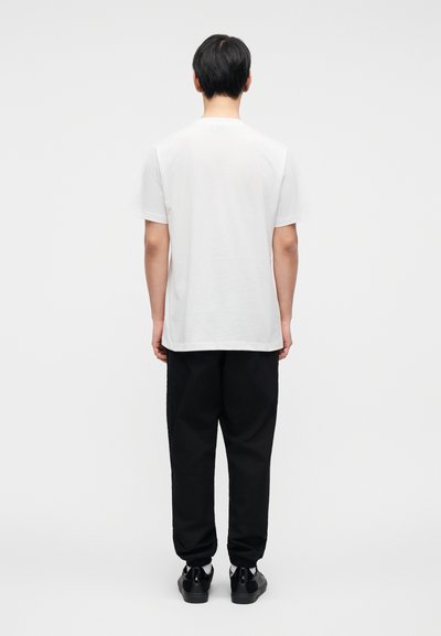 Λευκό βαμβακερό μπλουζάκι με στρογγυλή λαιμόκοψη, κοντά μανίκια και άνετη γραμμή, συνδυασμένο με μαύρο παντελόνι jogging και μαύρα παπούτσια.