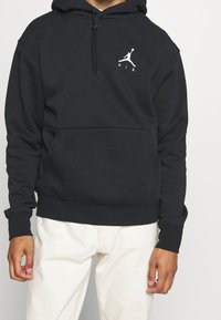 Uomo che indossa un hoodie nero con il logo Jumpman bianco e la scritta "AIR" sul petto, abbinato a pantaloni color avorio, in piedi davanti a uno sfondo semplice.