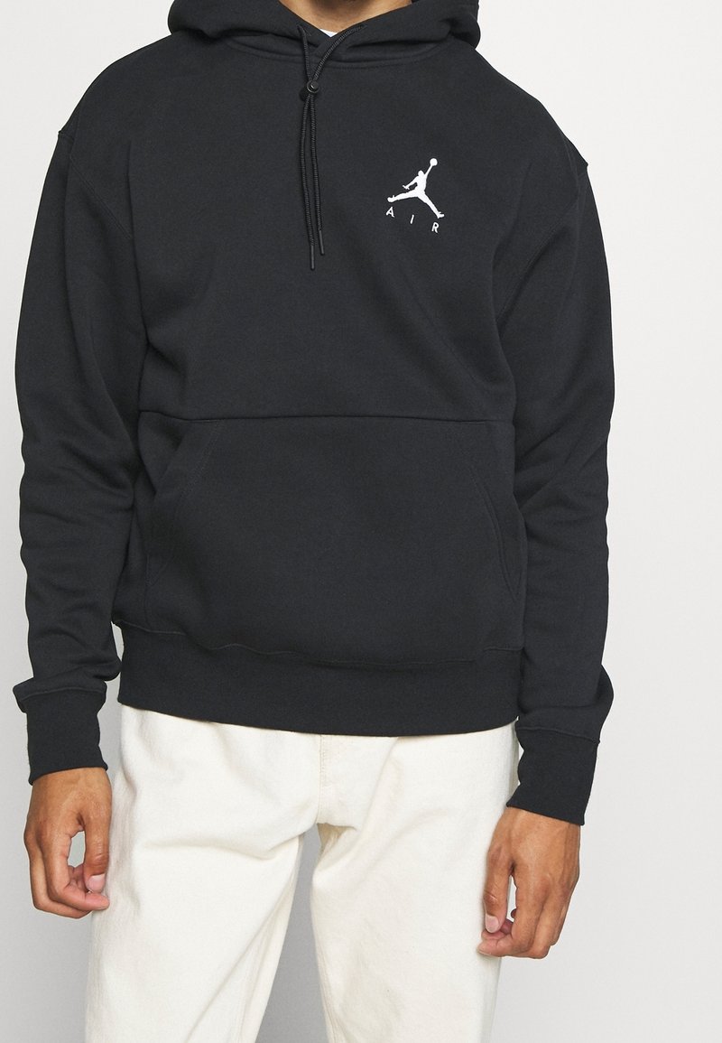 Uomo che indossa un hoodie nero con il logo Jumpman bianco e la scritta "AIR" sul petto, abbinato a pantaloni color avorio, in piedi davanti a uno sfondo semplice.