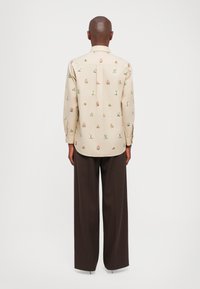 Lichtbeige overhemd met lange mouwen, voorzien van kleurrijke bloemenborduursels, een knoopsluiting kraag en plooidetail aan de achterkant. Gecombineerd met bruine wijde broek.