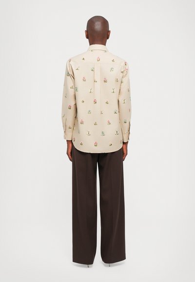 Jasnoszara długa koszula z długim rękawem z kolorowym haftem w kwiaty, z kołnierzykiem zapinanym na guziki i detalem plisowym z tyłu. W zestawie z brązowymi szerokimi spodniami.