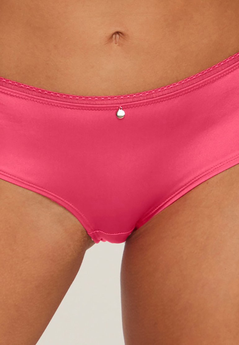 Slip bikini rosa con una texture liscia e lucida, caratterizzato da un dettaglio decorativo a forma di cuore argentato in vita e un bordo cucito a contrasto.