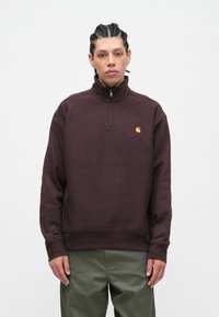 Sweat-shirt à col zippé quart en bordeaux, fabriqué en tissu doux. Dispose d'un petit logo jaune et de poignets côtelés. Convient pour une tenue décontractée.