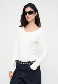 Λευκή μακρυμάνικη μπλούζα με ριμπ ύφανση και στρογγυλή λαιμόκοψη, συνδυασμένη με ψηλόμεσο σκουρόχρωμο υφασμάτινο τζιν και μαύρη ζώνη με τρουκς. Μαύρα γυαλιά ηλίου συμπληρώνουν το ντύσιμο.