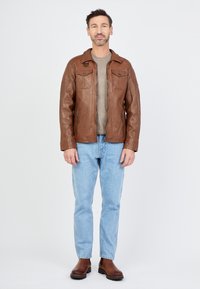 Braune Lederjacke mit Reißverschluss und zwei Brusttaschen, getragen über einem grauen Hemd, kombiniert mit hellblauen Jeans und braunen Stiefeln.