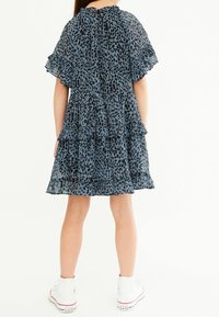 Vaaleansininen mekko, jossa on mustat leopardiäidit, röyhelöhihoja, kerroksellinen muotoilu ja pehmeä rakenne, yhdistettynä valkoisiin lenkkareihin.