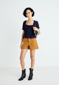Ung kvinne med kort, krøllete hår iført en marineblå topp med puff-ermer, sennepsgule shorts, svarte ankelstøvler og bærende en brun skulderveske.