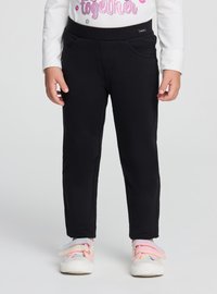 Bambino con pantaloni neri e scarpe da ginnastica color pastello, in piedi con le mani rilassate ai lati contro uno sfondo semplice.