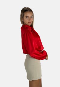Blusa rossa in satin con maniche lunghe e colletto a bottoni, abbinata a una gonna beige chiaro. Tessuto liscio e vestibilità ampia.